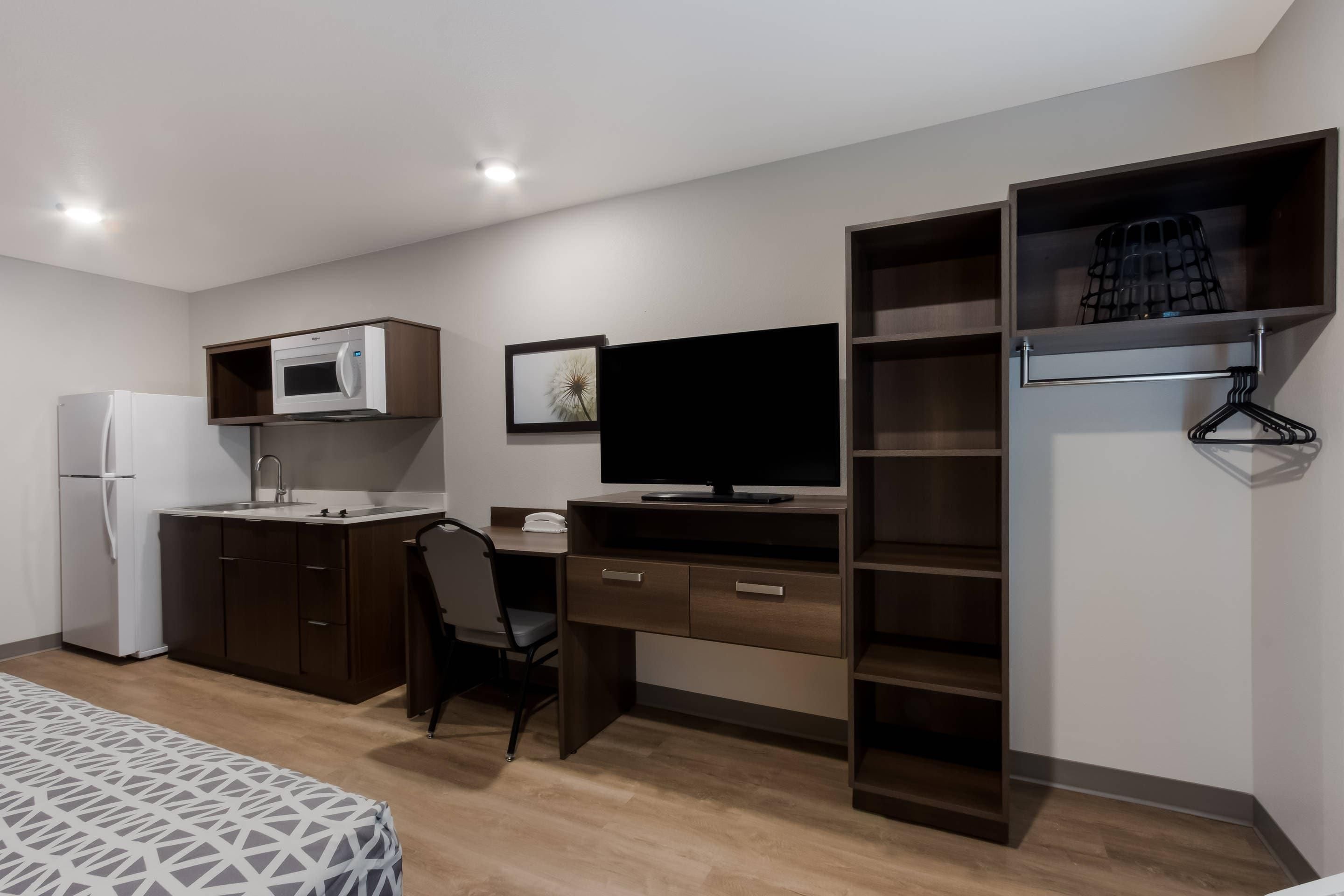 Quarto de hotel compacto com cozinha americana, cama com roupa de cama estampada, escrivaninha com cadeira, televisão e armário com cabideiro.