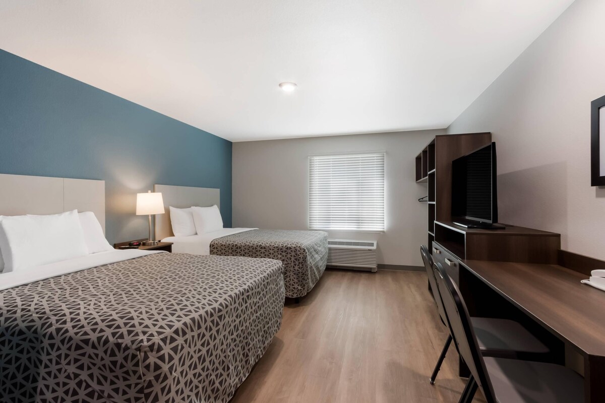 A hotel room with two beds, a desk, a TV, and a window with blinds.