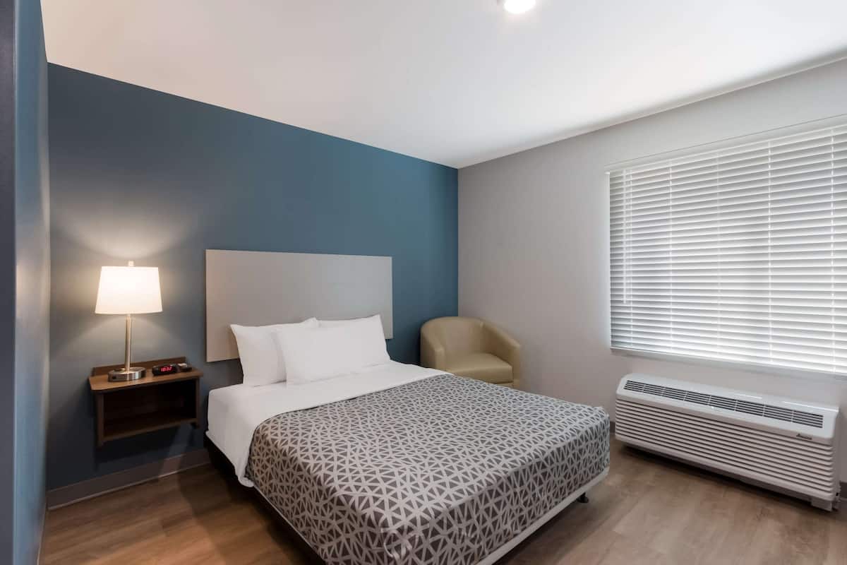 A hotel room with a bed, bedside lamp, chair, and air conditioning unit.