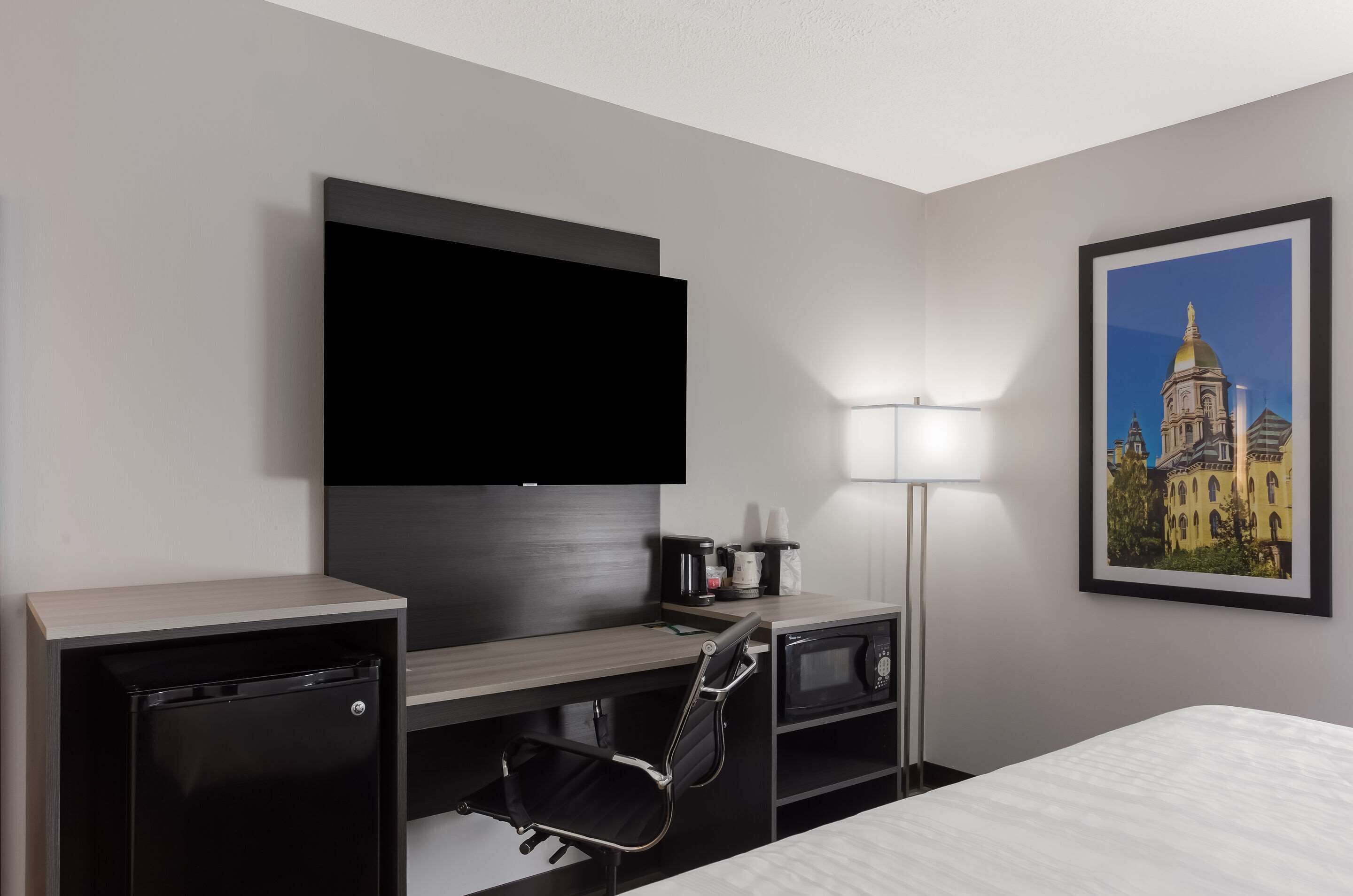 Habitación de hotel con cama, escritorio con televisor de pantalla plana, microondas, refrigerador pequeño, silla y lámpara.
