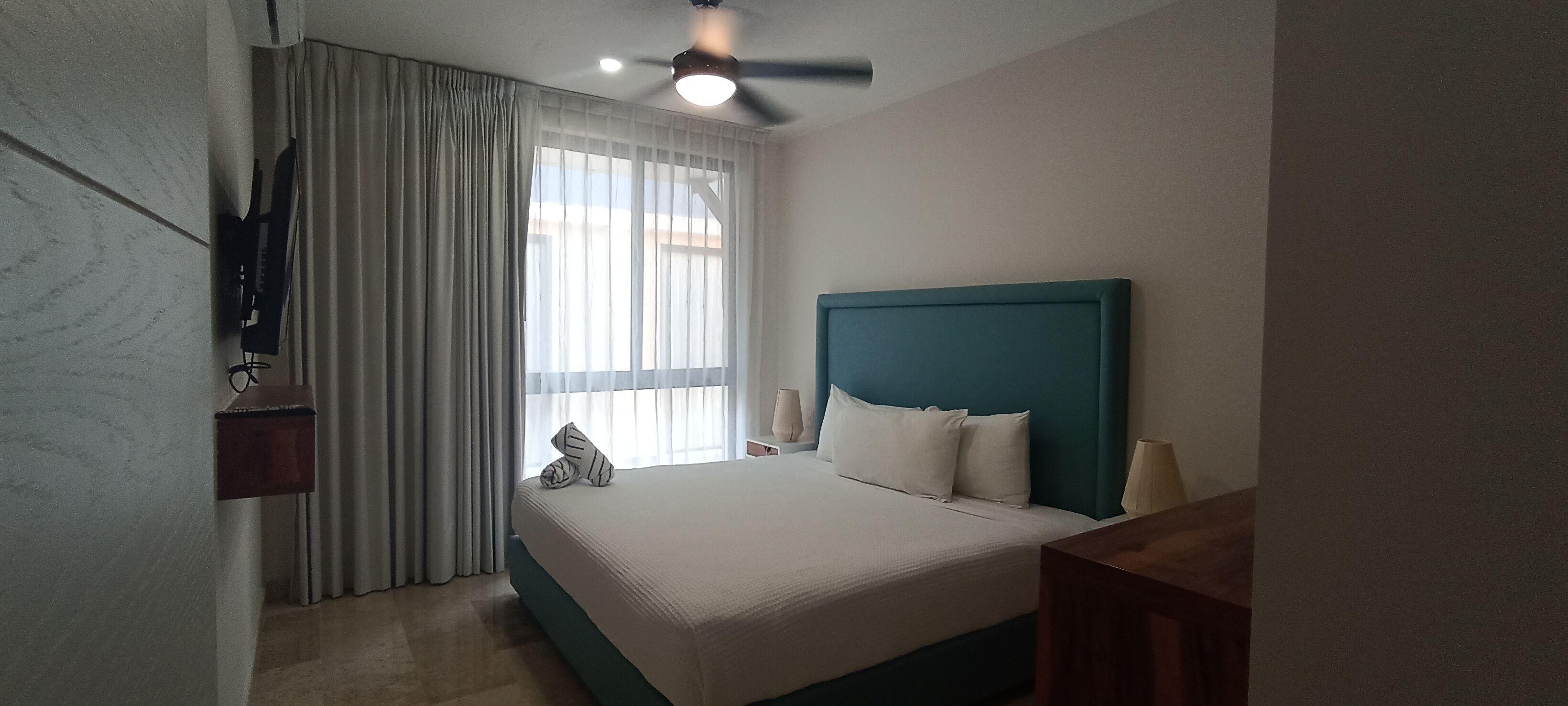 A hotel room with a bed, curtains, a ceiling fan, and a TV mounted on the wall.