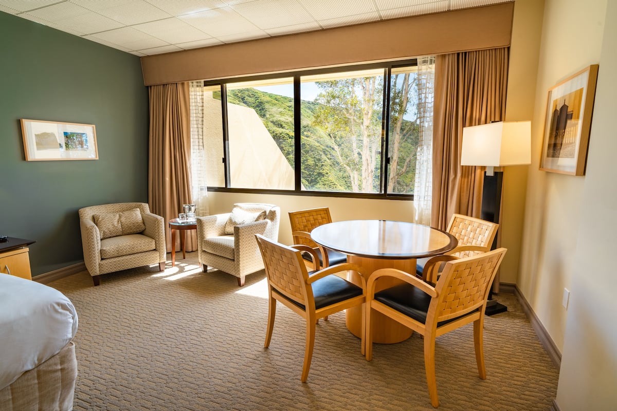 Habitación de hotel con un ventanal grande, una mesa redonda de comedor con sillas, un sofá, una mesita auxiliar y una lámpara.
