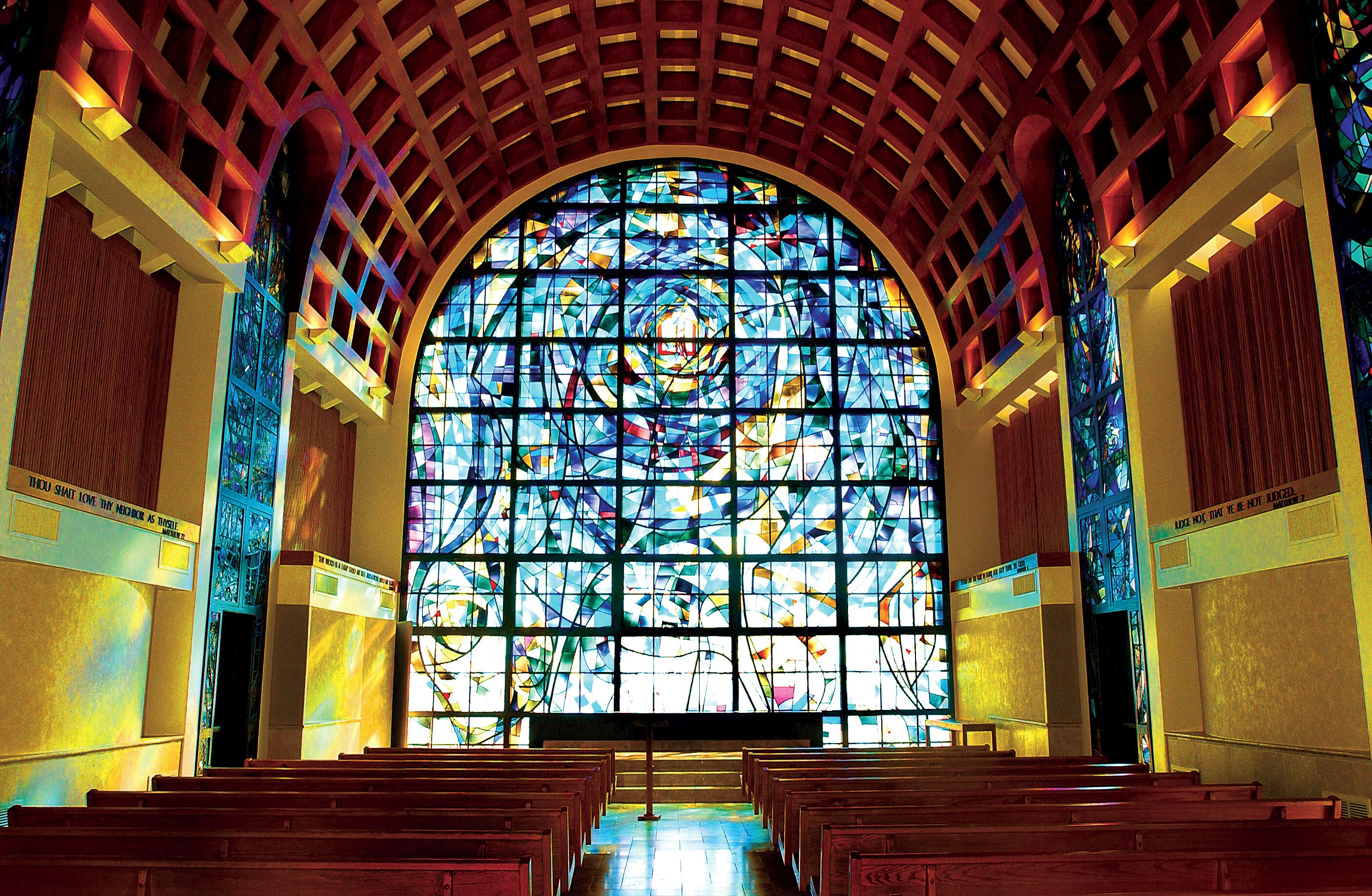 An interior with a large stained glass window, wooden pews, and a high arched ceiling.
