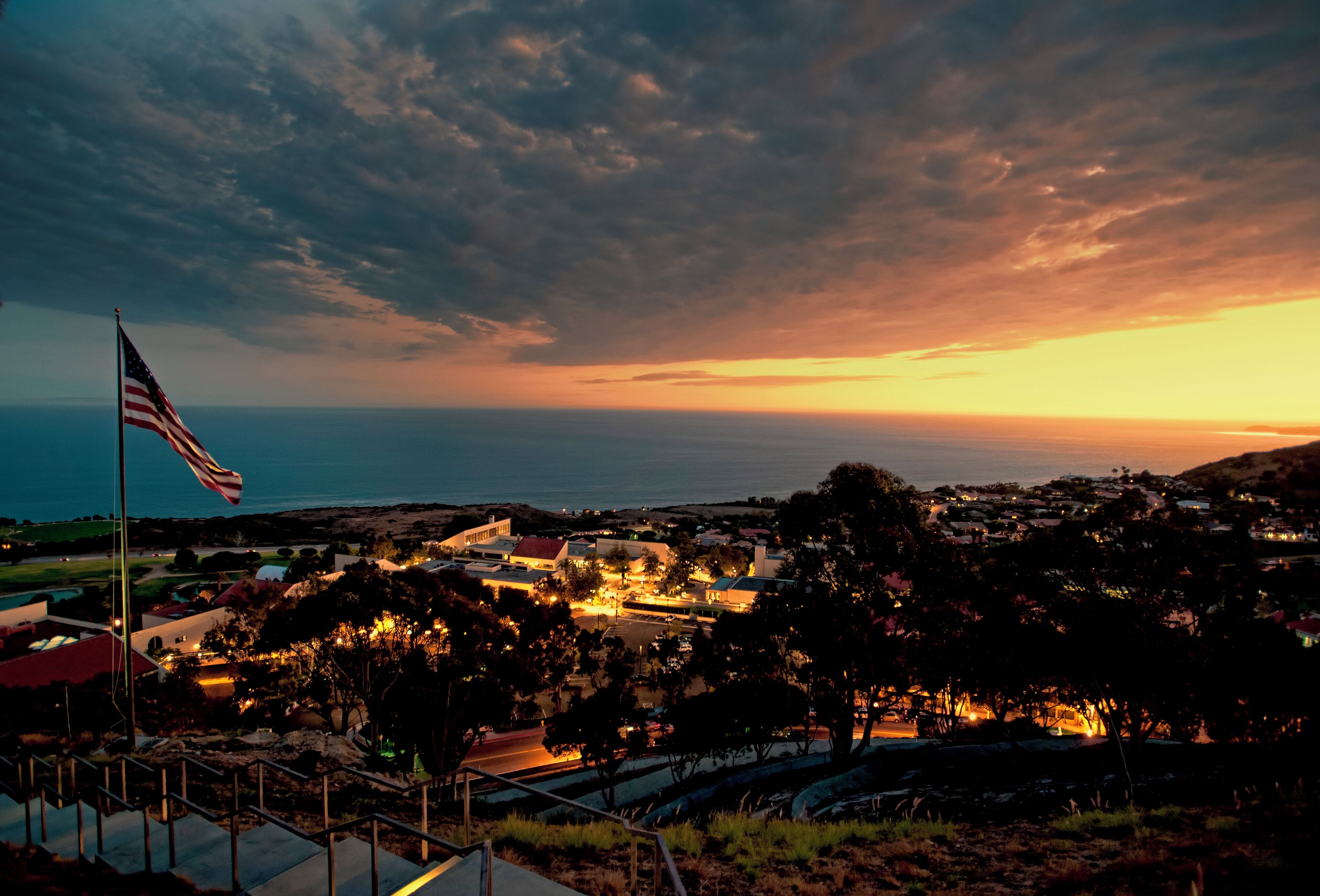 A coastal town at dusk with an American flag, illuminated buildings, and a view of the sea.