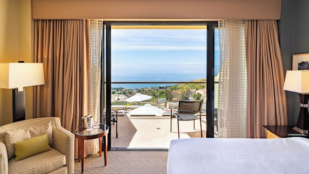 A hotel room with a bed, a chair, a small table, and a view of the ocean.