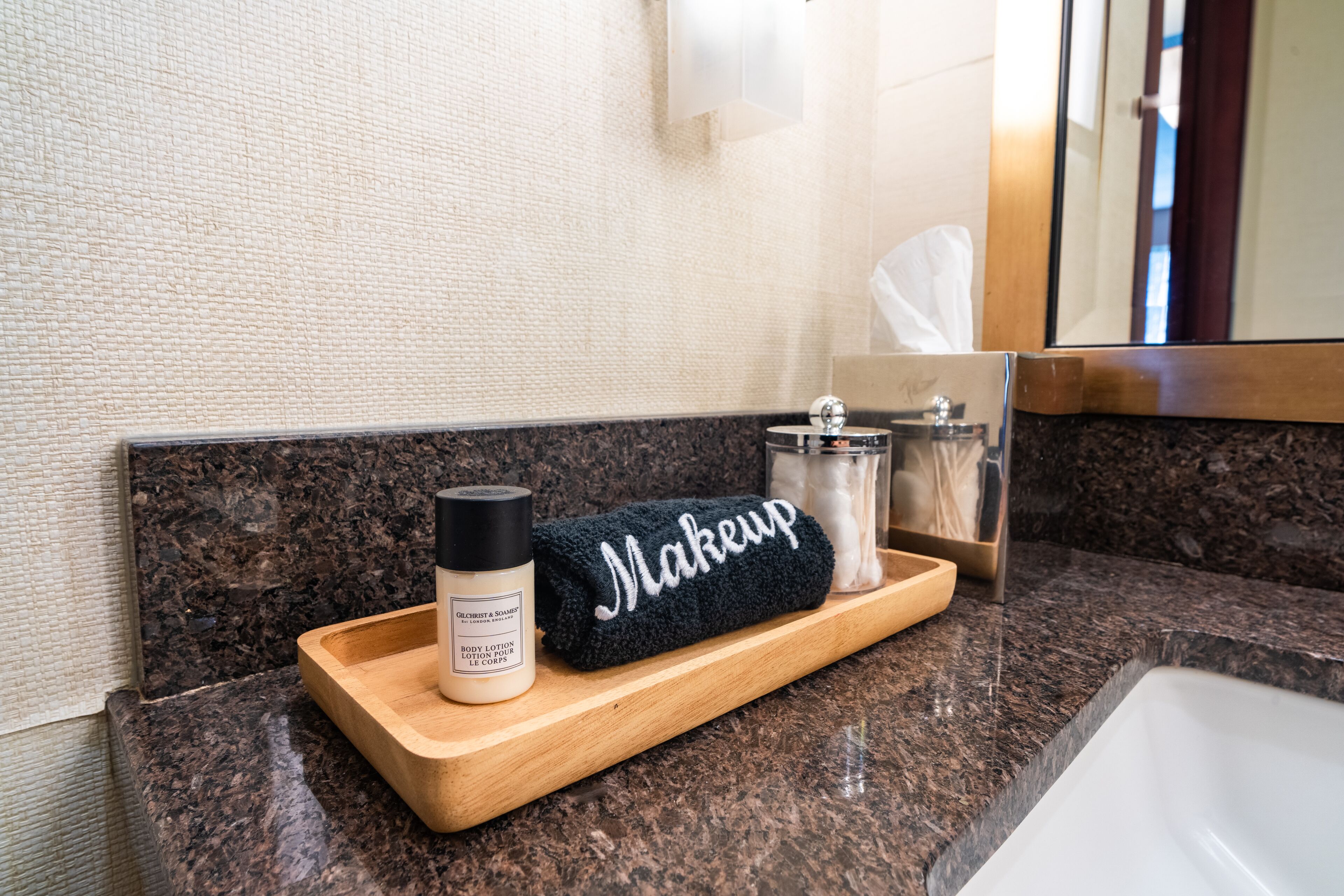 A bathroom vanity with a marble countertop, a wooden tray holding a black towel with the word "Makeup," a bottle of body lotion, and a glass jar with a lid.