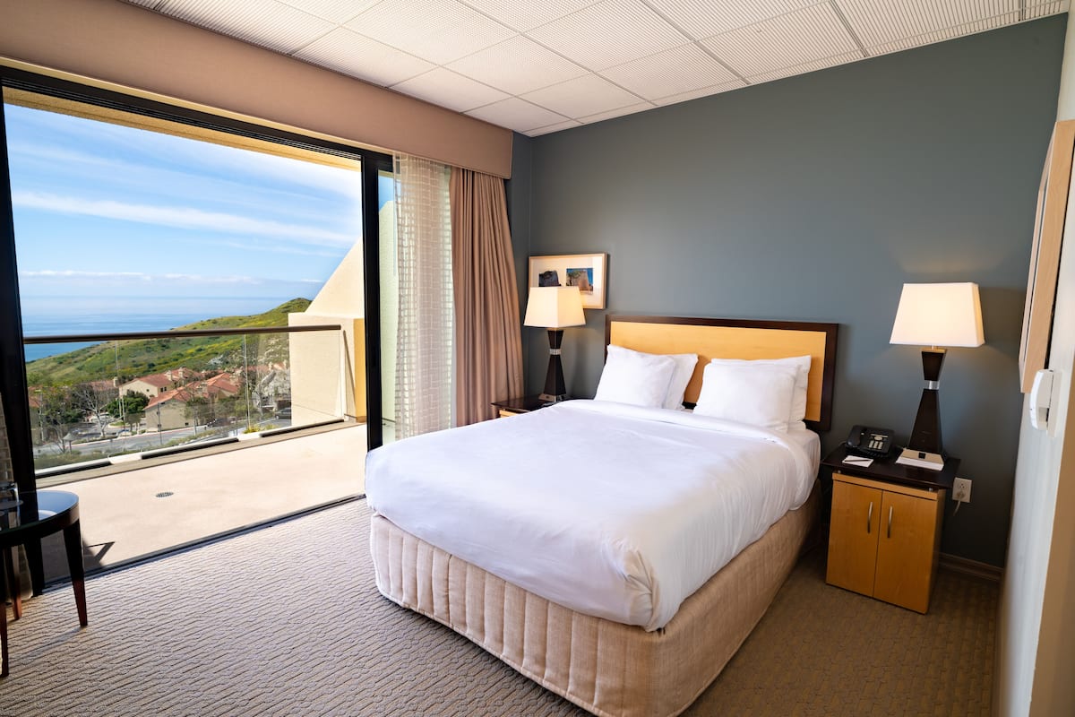 Habitación de hotel con una cama grande, dos mesitas de noche con lámparas, un escritorio pequeño y una silla. La habitación tiene un ventanal con vistas a un paisaje lejano.