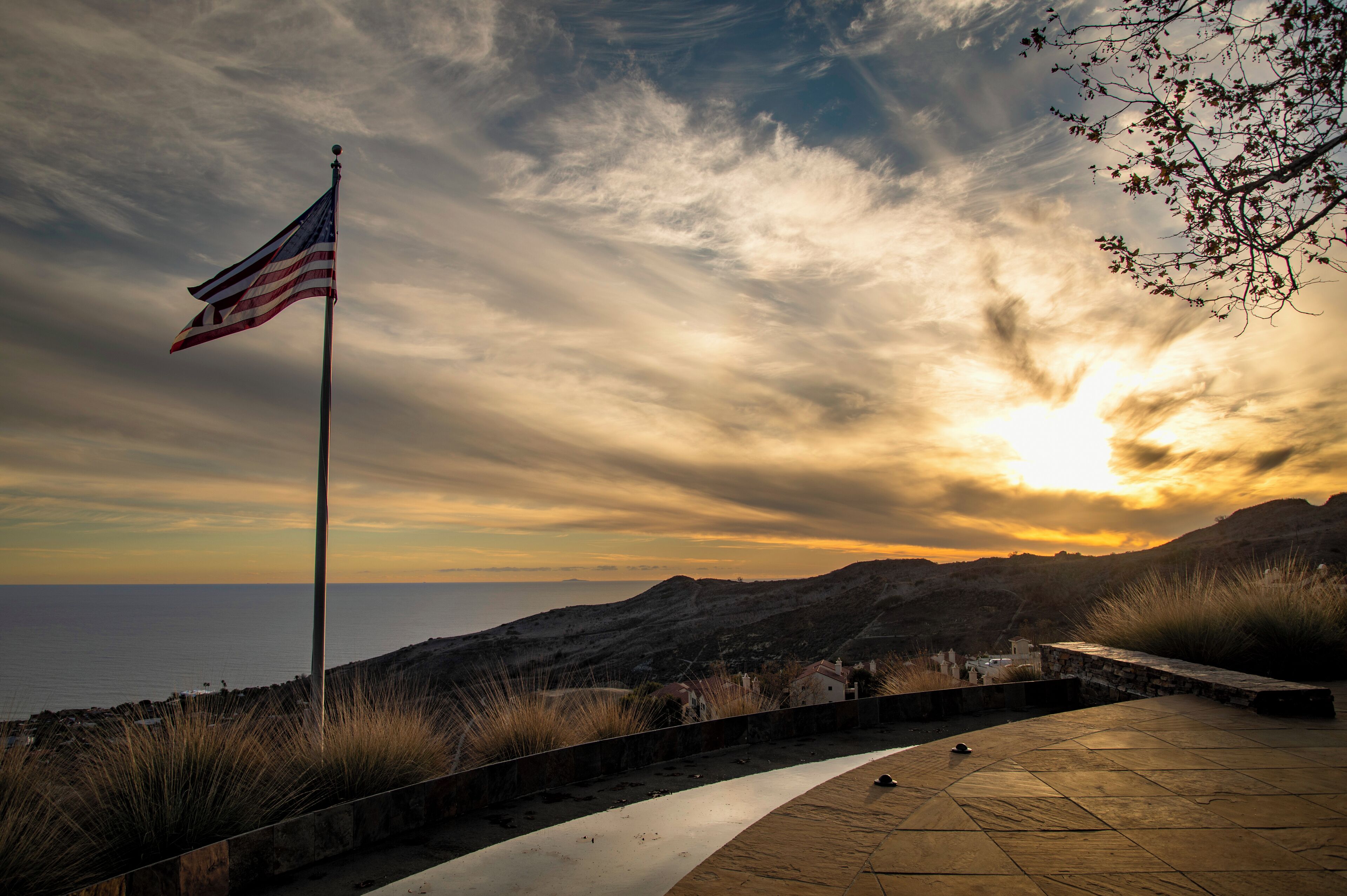 A flagpole with an American flag, a stone pathway, and a view of the sea and hills.