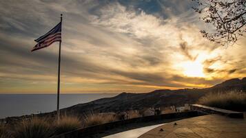 A flagpole with an American flag, a stone pathway, and a view of the sea and hills.