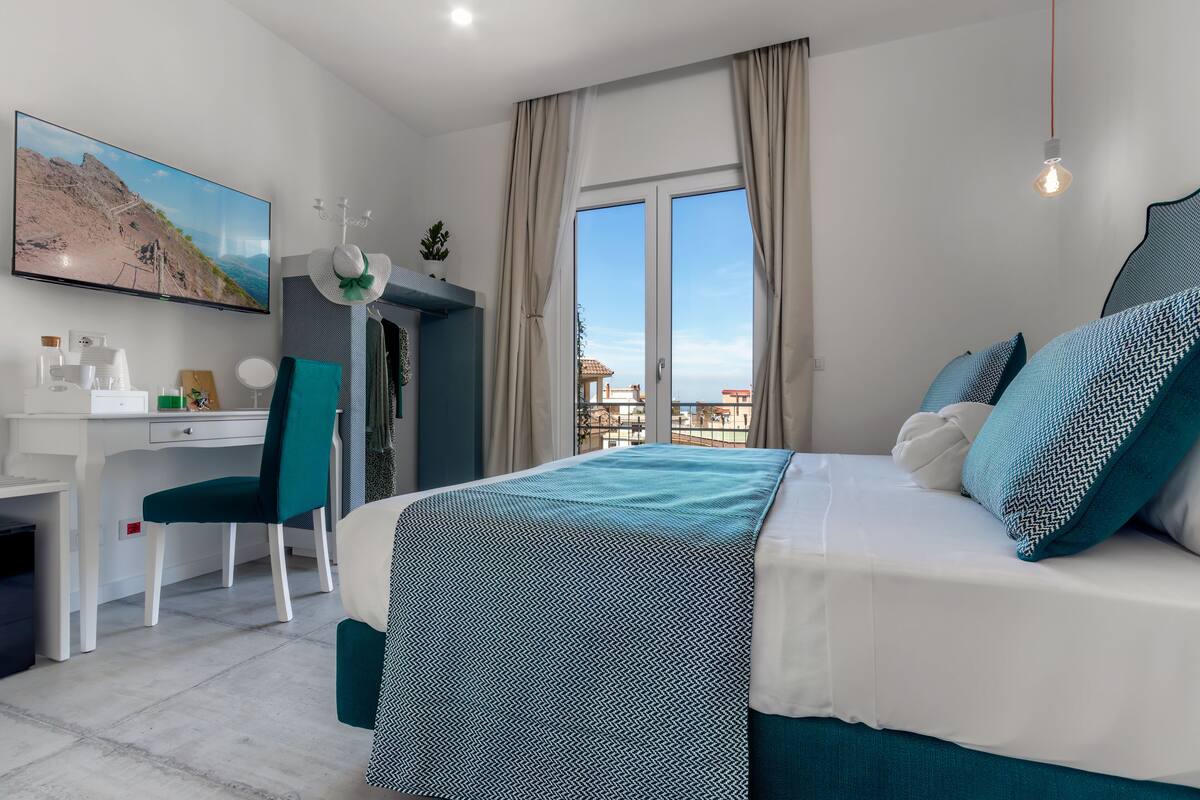 Ein Schlafzimmer mit Bett, Schreibtisch, Stuhl, Fernseher und einem Fenster mit Blick auf Gebäude.