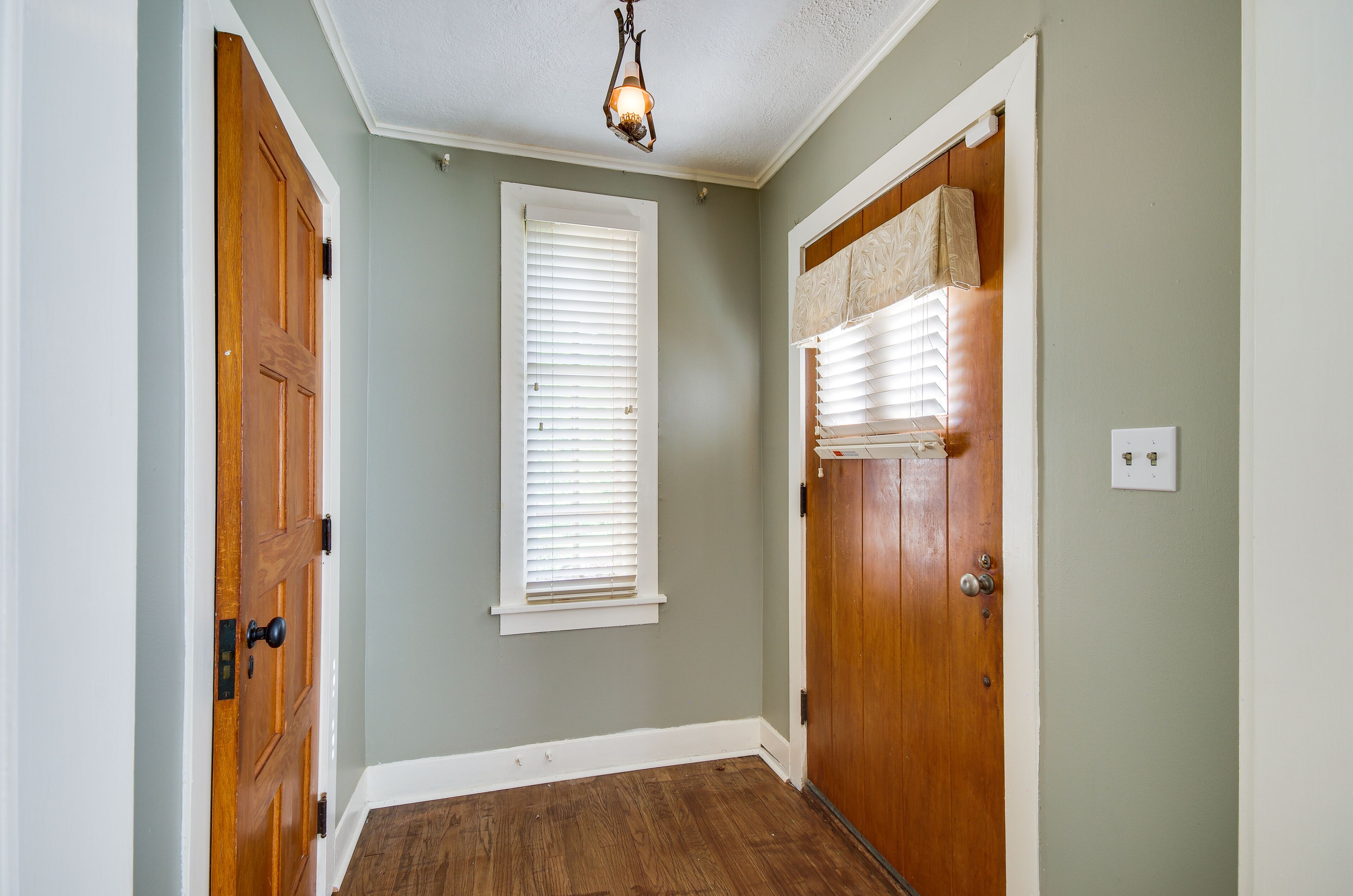 Un couloir avec des portes en bois, une fenêtre avec des stores et un luminaire au plafond.