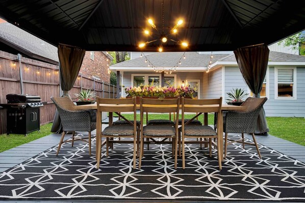An outdoor patio with a wooden table and chairs, a patterned rug, and string lights above.