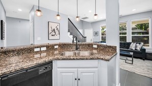 A modern kitchen with granite countertops, a stainless steel sink, and a dishwasher. There is a staircase leading to another level, and a black leather sofa in the adjacent living area.
