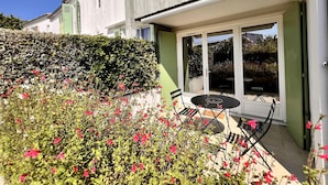 A modern house with a patio area featuring a table and chairs, surrounded by lush greenery and flowering plants. - Harmony, new apartment with heated pool (La Flotte)