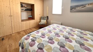 A bedroom with a wooden wardrobe, a bed with a colorful patterned bedspread, a chair, and a beach view painting. - Harmony, new apartment with heated pool (La Flotte)