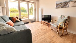 A living room with a grey sofa, a wicker chair, a flat-screen TV, and a large window with a view of greenery. - Harmony, new apartment with heated pool (La Flotte)