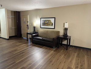 A hotel room with a brown leather sofa, two side tables with lamps, a framed picture on the wall, and a wooden floor. - Suburban Studios Winston-Salem near Hanes Mall (Winston-Salem)