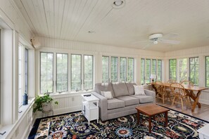 A sunroom with a gray sofa, wooden dining table, and chairs, a patterned rug, and large windows.