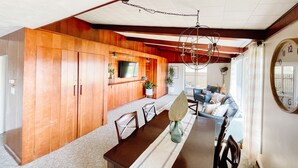 A dining area with a wooden table, chairs, and a chandelier. There is a television mounted on the wall and a clock on the wall.