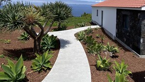 Un chemin menant à une maison dont le toit est recouvert de tuiles rouges, entourée de différentes plantes et offrant une vue sur la mer.