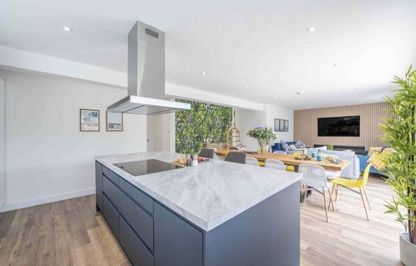 Een moderne keuken met een centraal eiland, een eethoek met een houten tafel en stoelen, en een woonkamer met een bank en televisie.