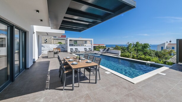 A modern outdoor pool area with a dining table and chairs, a covered seating area, and a view of the sea. - Villa Luna- Private Heated Pool (Novalja)