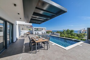 A modern outdoor pool area with a dining table and chairs, a covered seating area, and a view of the sea. - Villa Luna- Private Heated Pool (Novalja)