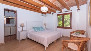 Une chambre à coucher avec un lit, des tables de chevet, une fenêtre, un coin repas avec des chaises en osier, et une cuisine visible à travers une porte ouverte.