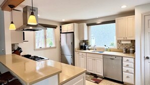 A modern kitchen with white cabinets, a stainless steel refrigerator, a dishwasher, and a large island with a sink. There are pendant lights above the island and a window providing natural light. - Path to Herring Pond Beach (Eastham)