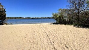 A sandy beach with a clear blue sky, a calm lake, and a small building with signs. - Path to Herring Pond Beach (Eastham)