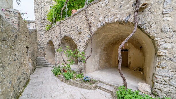 A narrow stone pathway leading to an arched doorway, flanked by stone walls and greenery.