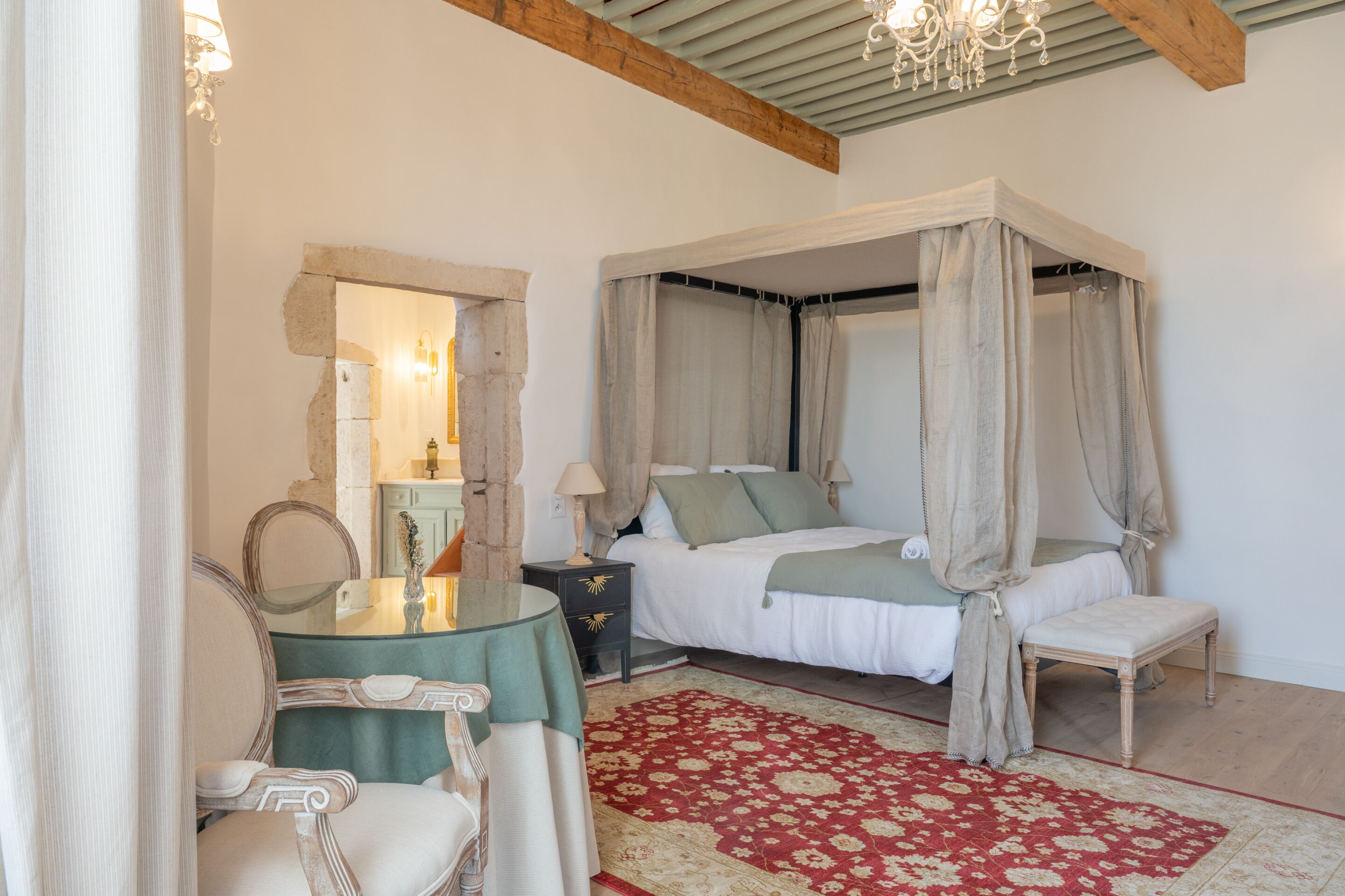 A bedroom with a four-poster bed, a green table with a chair, a red patterned rug, and a chandelier.
