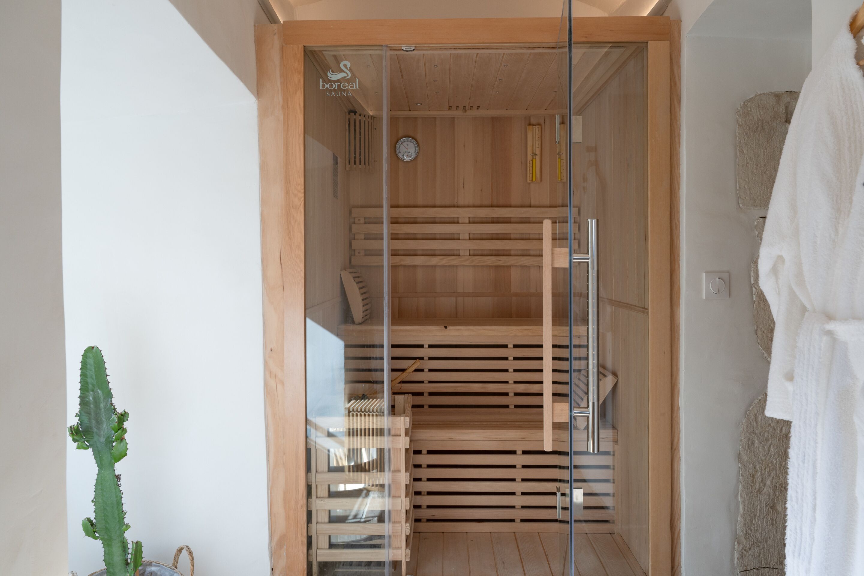 Een sauna met houten banken en een glazen deur, een klok aan de muur en een handdoekhouder aan de rechterkant.