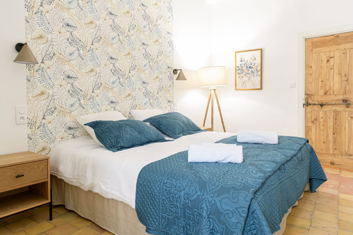 A neatly made bed with a blue bedspread and pillows, a bedside lamp, a wooden nightstand, and a patterned wallpaper.