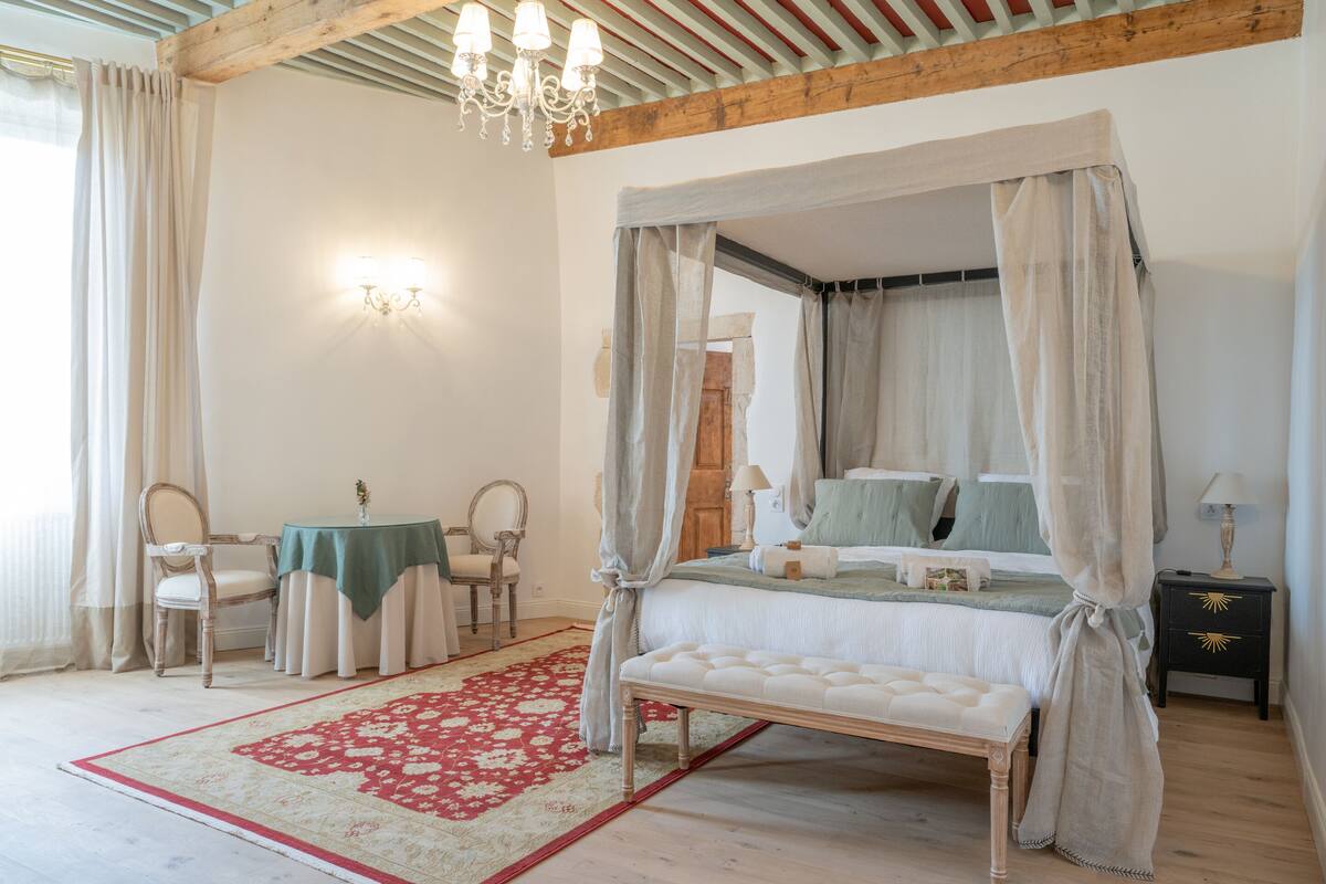 A bedroom with a four-poster bed, a red and white patterned rug, a wooden bench, a nightstand with a lamp, and a table with chairs.