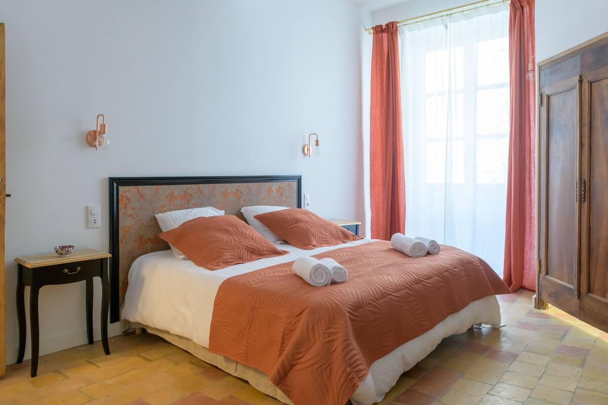 A neatly made bed with a matching headboard, a bedside table with a bowl, and a window with curtains.