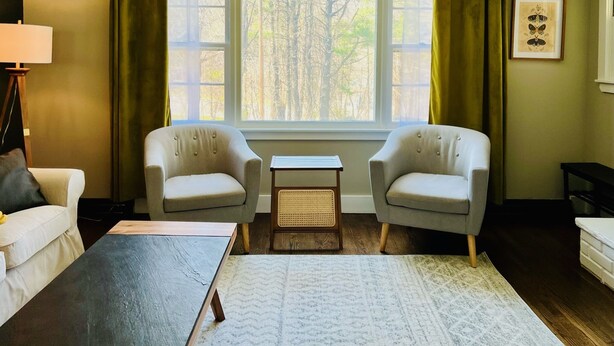 A living room with two armchairs, a small table, and a couch. There is a window with curtains, a lamp, and a framed picture on the wall. - Appaltree Bungalow: Two bedrooms in town (Boone)
