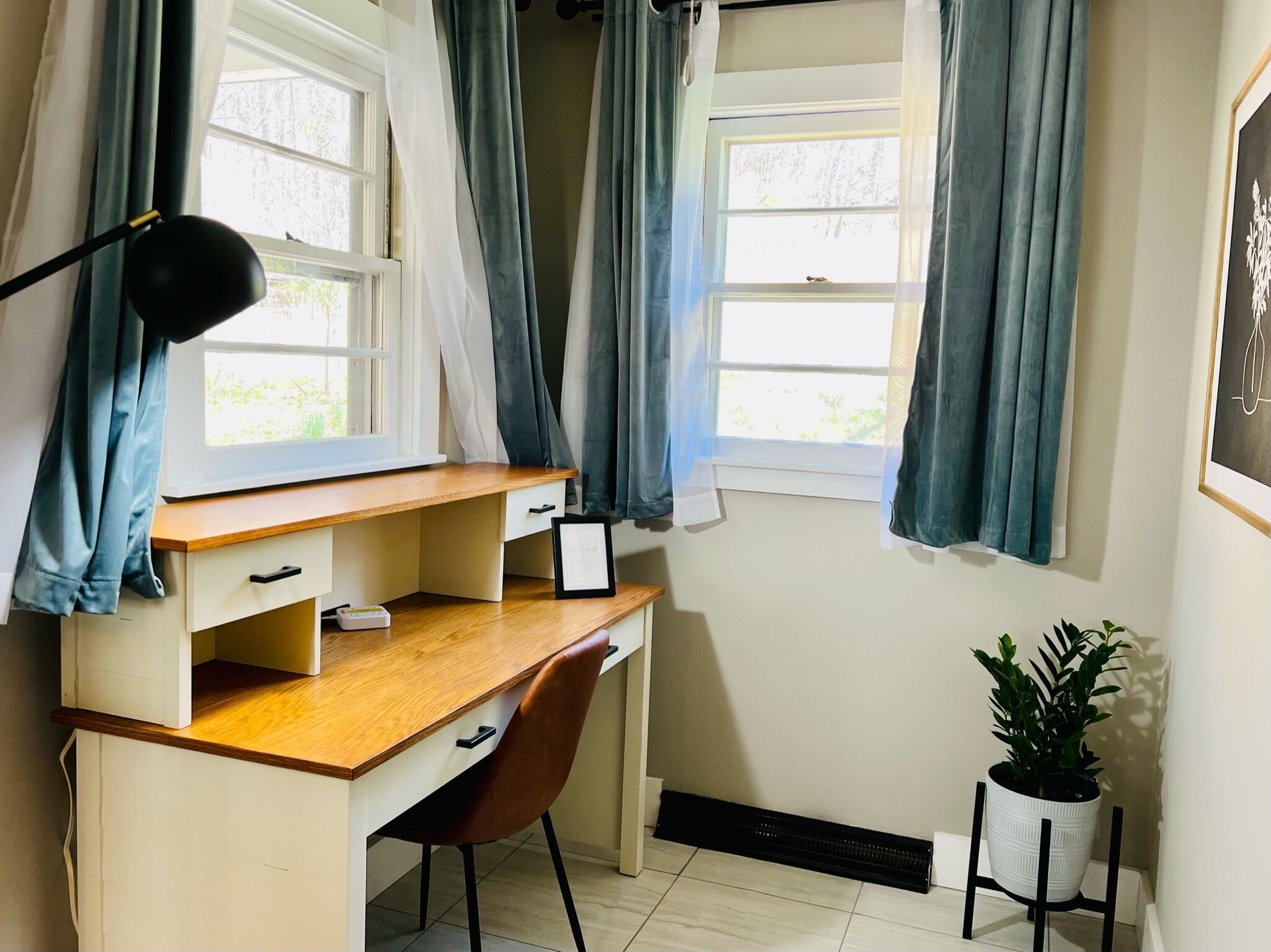 Un estudio con escritorio de madera, dos ventanas con cortinas, lámpara de escritorio, una silla, radiador y una planta en maceta.