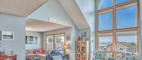 A spacious living room with a high ceiling, large windows offering a view of the sea, a cozy seating area with a blue armchair and a bookshelf filled with books and decorative items.
