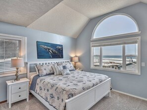 A bedroom with a bed, two bedside tables, a wall-mounted lamp, a painting, and a large window with a view.