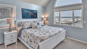 Un dormitorio con cama, mesitas de noche, un cuadro de ballenas, un ventanal con vista al océano y un techo inclinado.