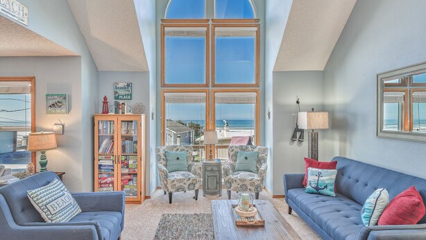 Amplia sala de estar con techo alto, ventanas grandes que ofrecen vista al océano, un rincón de descanso acogedor con sofá y sillones azules, y una estantería llena de libros y objetos decorativos.
