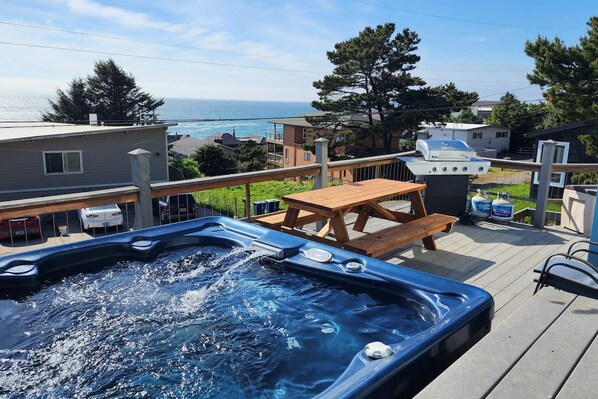 A hot tub on a deck with a view of the ocean, a picnic table, and a barbecue grill.