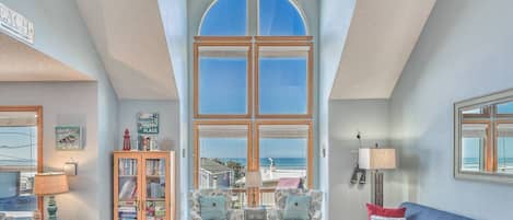 A spacious living room with a high ceiling, large windows, and a view of the sea.