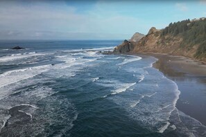 Un paisaje costero con olas rompiendo sobre una playa arenosa, un acantilado rocoso y una ladera cubierta de bosque.