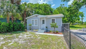 Ein eingeschossiges Haus mit Veranda, umgeben von einem Maschendrahtzaun, und ein kleiner Garten mit spärlichem Gras und einigen Bäumen.