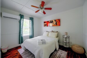 A bedroom with a bed, a ceiling fan, a painting, a bedside table with a lamp, and a window with curtains. - Pet-Friendly Cottage Near Beach: King Bed/Fire Pit (Bonita Springs)