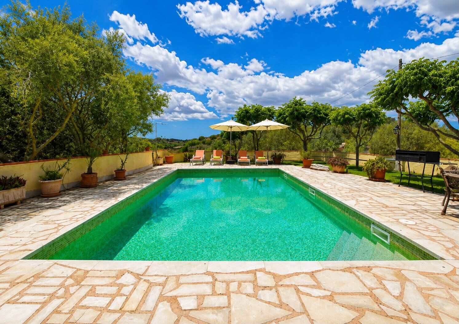 Una piscina rectangular amb un marc de pedra, envoltada de plantes en testos i mobiliari exterior.