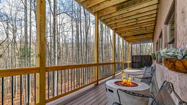 Une terrasse extérieure couverte, dotée d’un foyer et d’un espace salon, entourée d’une rambarde en bois et offrant une vue sur une zone boisée.