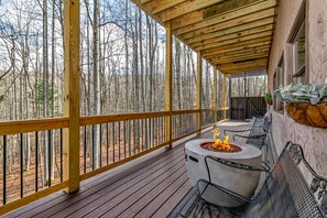 Terraza techada al aire libre con un brasero y zona de estar, rodeada por barandilla de madera y con vistas a un área boscosa.